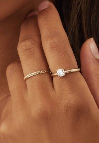 Des bagues en or sur une main : l'une est un anneau tressé, et l'autre présente une pierre ronde et claire sertie dans un anneau en or simple.