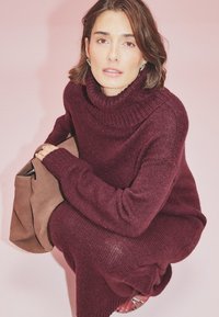 Burgundia värvi kõrge kaelusega kampsun, mis on valmistatud pehmest kudumaterjalist, omades lõdvemat lõiget ja soonikute detaile. kõrval lebab neutraalne beež käekott.