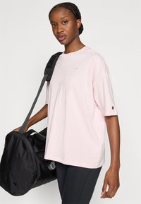 Ροζ ανοιχτό ροζ κοντομάνικο μπλουζάκι με στρογγυλή λαιμόκοψη και μικρό λογότυπο στο στήθος. Μαύρη τσάντα duffel με λουρί ώμου και υφασμένο χερούλι.