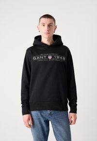 Felpa nera con cappuccio e logo GANT 1949 in bianco e rosso. Realizzata in materiale morbido, presenta una tasca a canguro e polsini a coste.