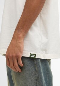 Valge puuvillane T-särk, millel on lõdvestunud lõige, lühikesed varrukad ja roheline Barbour silt vasakul küljel hems, koos heledate denim pükstega.