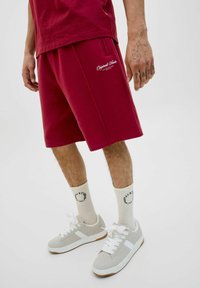 Shorts en coton bordeaux avec poches latérales et détail de logo, associés à des baskets gris clair et des chaussettes crème avec texte noir.