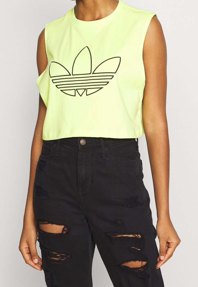 Top corto sin mangas de neón amarillo con logo negro de Adidas. Combinado con jeans negros desgastados con rasgaduras y bordes deshilachados.