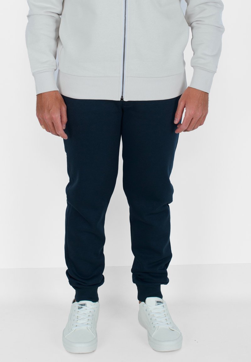 Felpa leggera grigia con zip, maniche e orlo a coste, abbinata a pantaloni da jogging blu navy e sneakers bianche, caratterizzata da una texture liscia e un design aderente.