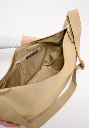 Beige Umhängetasche, offen gehalten, um das große Hauptfach und das innere Reißverschlussfach mit dem Aufkleber „FROM TRASH TO TREASURE“ zu zeigen.