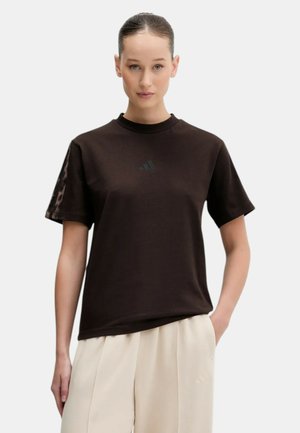 Giovane donna con pelle chiara e capelli raccolti in una coda di cavallo, indossa una maglietta nera Adidas e pantaloni beige, in piedi davanti a uno sfondo semplice.