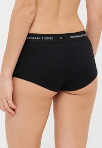Svart ribbestrikkede bomullstruser med bred elastisk midjebånd som har hvit tekst "Copenhagen Studios," og har en tettsittende design og glatt tekstur.