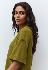 Olivengroen oversized T-shirt met een voorzak, ongezoomde mouwen en een zachte, getextureerde stof. Model met lang donker haar, zijwaarts gericht.