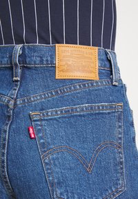 Modré denimové džíny s béžovou koženou nášivkou, na které je napsáno "Levi Strauss & Co." Zlaté prošívání a malá červená nášivka na levé kapse.