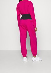 Fuchsia träningsoverall med svart elastiskt midjeband, långa ärmar och smalnande byxor, matchad med vita sneakers. Har en bakfickadetalj.