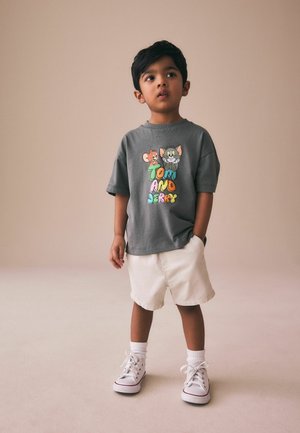 Fiatal fiú áll, szürke Tom és Jerry pólót, bézs rövidnadrágot, fehér zoknit és fehér tornacipőt visel, kissé oldalra néz.
