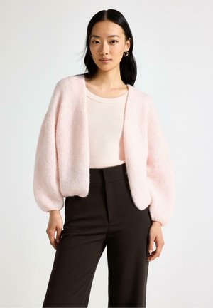 Donna che indossa un cardigan peloso rosa chiaro sopra una maglia rosa pallido e pantaloni neri a vita alta, in piedi contro uno sfondo chiaro e semplice.