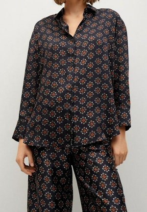 Kobieta ubrana w czarną koszulę zapinaną na guziki i spodnie z powtarzającym się czerwono-białym geometrycznym wzorem kwiatowym.