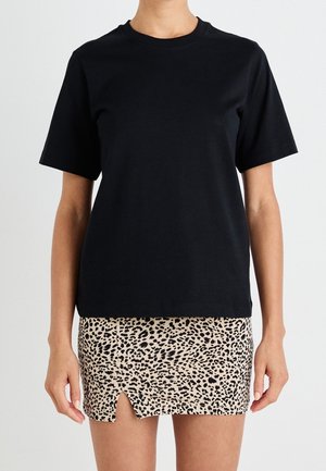 Kvinde iført en ensfarvet sort T-shirt med korte ærmer, kombineret med en beige mininederdel med sort leopardprint og frontslids.