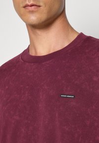 Vyno raudona trumpa rankovė t-shirt, pagaminta iš minkštos medvilnės, su apvalia iškirpte ir mažu juodu "UNDER ARMOUR" logotipu priekyje.