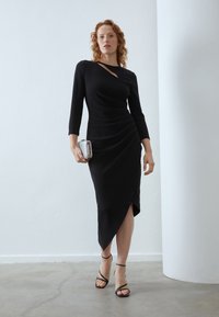 Schwarzes Bodycon-Kleid mit langen Ärmeln, asymmetrischem Cut-Out-Detail, drapiertem Textur und einem seitlichen Schlitz. Kombiniert mit einer kleinen silbernen Clutch und Riemchenschuhen.