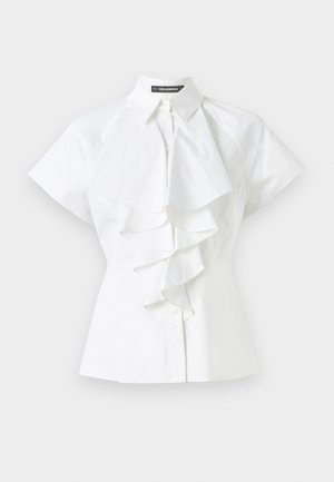 Balta blūze ar apkakles izgriezumu un īsām piedurknēm. Rūtainas detaļas iet gar centru, izgatavota no gluda kokvilnas auduma.