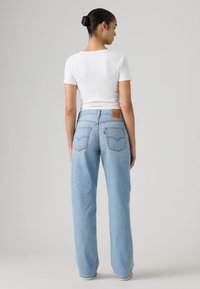 Γυναίκα με σκούρα μαλλιά πιασμένα σε κότσο, που φοράει λευκό τοπ με κοντά μανίκια και ανοιχτόχρωμο τζιν ψηλόμεσο, στέκεται γυρισμένη πλάτη σε απλό φόντο.