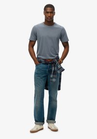 Grijze korte mouwen crewneck t-shirt, blauwe spijkerbroek, geruit overhemd om de taille geknoopt, beige sneakers met gestructureerde details.