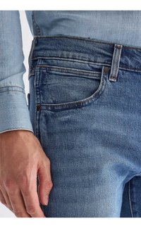 Közeli kép egy személyről, aki közép kék farmert és világoskék farmeringet visel, a bal kezével az oldalán pihenve.