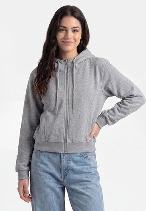 Frau lächelt, trägt eine hellgraue Kapuzenjacke mit Reißverschluss und Kordeln sowie eine hellblaue Jeans und posiert mit einer Hand in der Hüfte vor einem schlichten Hintergrund.