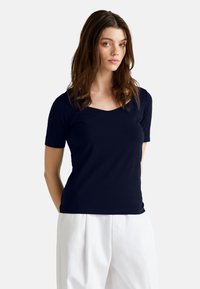 Námořnická modrá tričko s krátkým rukávem a výstřihem do V. Vyrobeno z měkké tkaniny s hladkým povrchem. Nosené s bílými kalhotami s vysokým pasem.