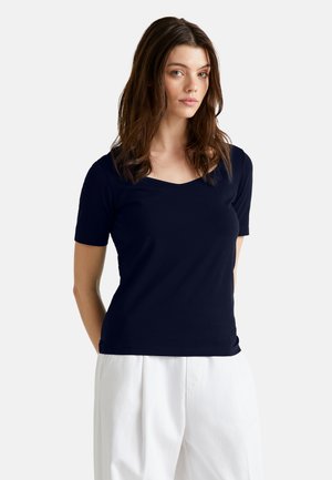 Marineblaues T-Shirt mit kurzen Ärmeln und V-Ausschnitt. Hergestellt aus weichem Stoff mit glatter Textur. Kombiniert mit weißen hoch taillierten Hosen.