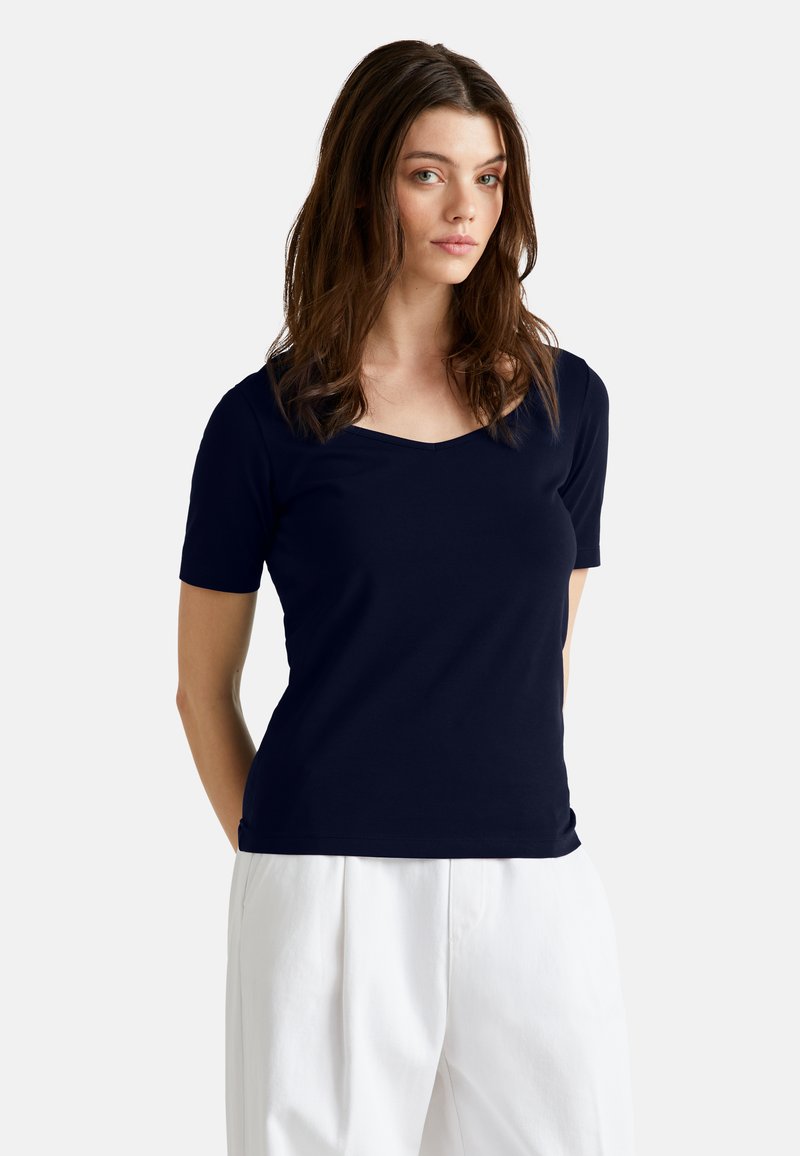 Námořnická modrá tričko s krátkým rukávem a výstřihem do V. Vyrobeno z měkké tkaniny s hladkým povrchem. Nosené s bílými kalhotami s vysokým pasem.