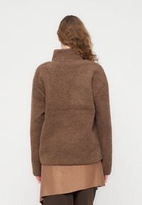 Καφέ φλις που λόβερ με ψηλό γιακά, υφασμάτινη επιφάνεια με υφή και μακριά μανίκια, σχεδιασμένο για ζεστασιά και άνεση. Φοριέται πάνω από καφέ μπλουζάκι.