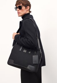 Musta viestintälaukku, joka on valmistettu teksturoituneesta kankaasta ja siinä on nahkaisia yksityiskohtia. Laukussa on logo, säädettävä olkahihna ja siihen on kiinnitetty pieni tagi.