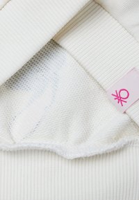Indumento bianco caratterizzato da una texture a coste e una fodera interna liscia. Include un piccolo etichetta rosa con un dettaglio grafico su un lato.