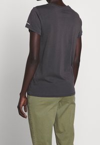 Tmavě šedé bavlněné tričko s krátkým rukávem a kulatým výstřihem, zobrazené zezadu. Pozoruhodný detail loga na levém rameni. Olivově zelené kalhoty.