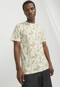Camiseta de algodón de color claro con un patrón floral en beige y blanco, de cuello redondo y mangas cortas. Llevada con pantalones cortos de mezclilla oscuros.