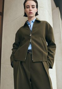 Oliivinvihreä bomber-takki vetoketjulla, isoilla taskuilla ja vaaleansininen paita alla. Asun täydentävät räätälöidyt housut.