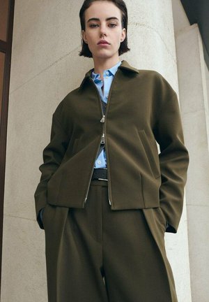 Veste bomber vert olive avec fermeture éclair, grandes poches, et une chemise bleu clair en dessous. Un pantalon sur mesure complète la tenue.