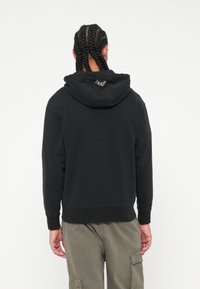 Μαύρη hoodie με τσέπη καγκουρό, ριμπ μανσέτες και στρίφωμα. Διαθέτει λογότυπο ραμμένο με μηχανή στη πλάτη. Λείος βαμβακερός ύφασμα.
