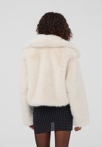 Giacca in pelliccia sintetica color crema con ampio colletto, maniche lunghe e una texture morbida. Abbinata a una gonna nera con pois bianchi.