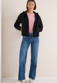 Mujer vestida con una sudadera negra con cremallera, camiseta rosa, jeans azules y zapatillas blancas, de pie contra una pared beige con las manos en los bolsillos.