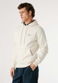 Beige hoodie met aantrekkoord, zak aan de voorkant en logo. Zachte stof, ribgebreide manchetten en zoom. Gecombineerd met donkere broek.