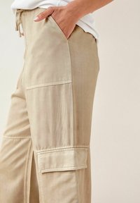 Beige, lette cargo-bukser med en sidelomme, snore i taljen og glat tekstur. Enkel design med minimale syninger.