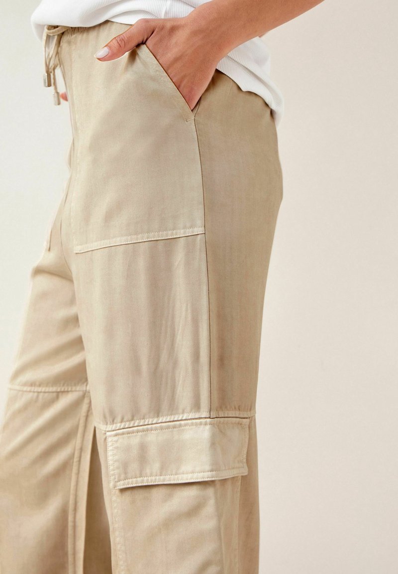 Beige, lette cargo-bukser med en sidelomme, snore i taljen og glat tekstur. Enkel design med minimale syninger.