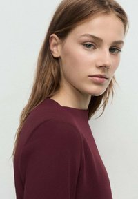 Top carmesí con un cuello alto y líneas limpias, que presenta una textura suave y un diseño minimalista, llevado por un modelo con cabello castaño claro.