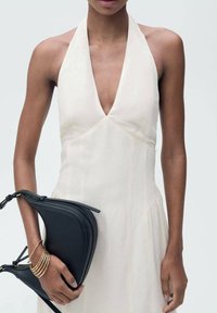 Abito crema con scollatura a V profonda e allacciatura al collo, caratterizzato da una silhouette fluida. La modella tiene in mano una borsa nera curva e braccialetti dorati.