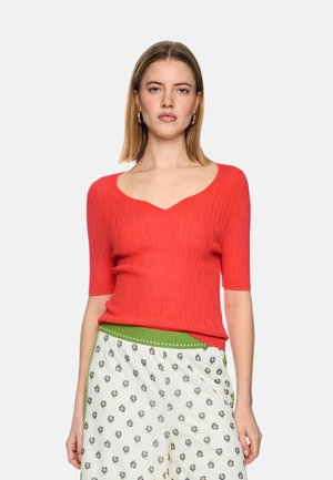 Frau mit schulterlangem blondem Haar, die ein rotes kurzärmeliges Oberteil und weiße, gemusterte Hosen mit grünem Bund trägt, steht vor einem einfarbigen Hintergrund.