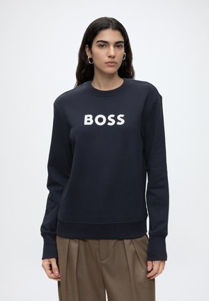 Femme aux longs cheveux foncés portant des boucles d'oreilles créoles dorées, un sweat-shirt bleu marine marqué BOSS et un pantalon plissé marron sur un fond uni.