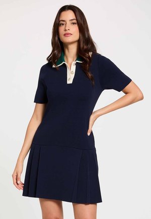 Mujer con un vestido azul marino de manga corta con falda plisada y cuello en contraste verde y beige con botones, posando con una mano en la cadera.