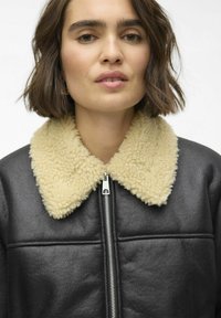 Veste en cuir noir avec un col en fausse fourrure beige. Possède une fermeture éclair à l'avant et des coutures horizontales sur la poitrine.