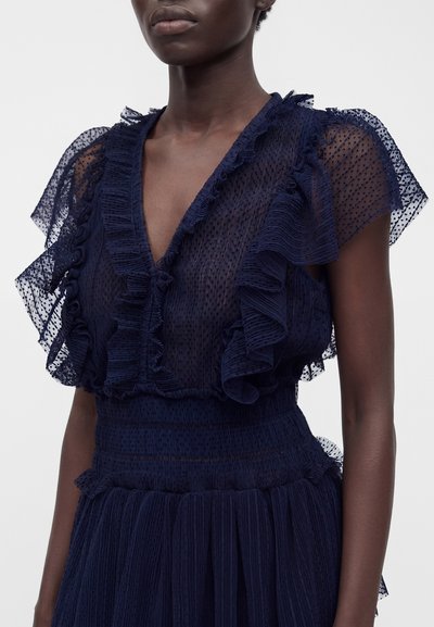 Personne à la peau foncée portant une robe bleu marine transparente avec des manches à volants, un décolleté en V profond et un tissu plissé texturé.