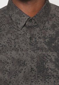 Chemise à boutons en gris foncé avec un motif noir tacheté. Dotée d'un col pointu et de boutons noirs, confectionnée en tissu texturé.