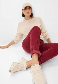 Beige gebreide trui, rode corduroy broek opgerold bij de enkels, en lichtbeige sneakers met textuuraccenten en veters. Neutrale achtergrond.